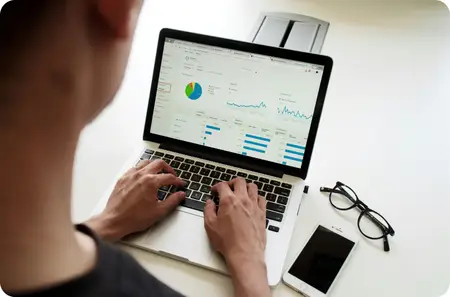 A professional working on a laptop displaying financial data, charts, and business analytics. The workspace is organized with a smartphone and eyeglasses placed nearby, creating a focused and modern setting.