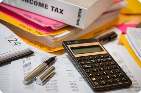 image showing a calculator, two pens, a printed spreadsheet, and a stack of documents, with a visible "INCOME TAX" book labeled "30th Edition" on top. A desk calendar displaying the number 24 suggests a tax deadline or important date.