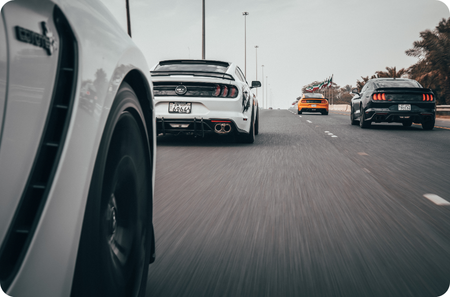 photograph captures a dynamic scene of multiple muscle cars speeding down a highway. The perspective is taken from the side of one vehicle, showing the rear view of a white Ford Mustang with a spoiler and custom license plate in the center. To the right, another black Mustang follows closely, while additional vehicles can be seen further ahead. The motion blur on the road and wheels enhances the sense of speed and movement.