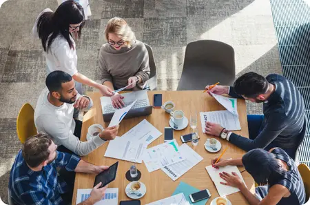 A group of six diverse professionals engaged in a collaborative meeting around a wooden table in a modern office setting. They are reviewing documents, using laptops and tablets, and discussing ideas while having coffee. The image is taken from an overhead perspective, capturing an energetic and engaged work environment.